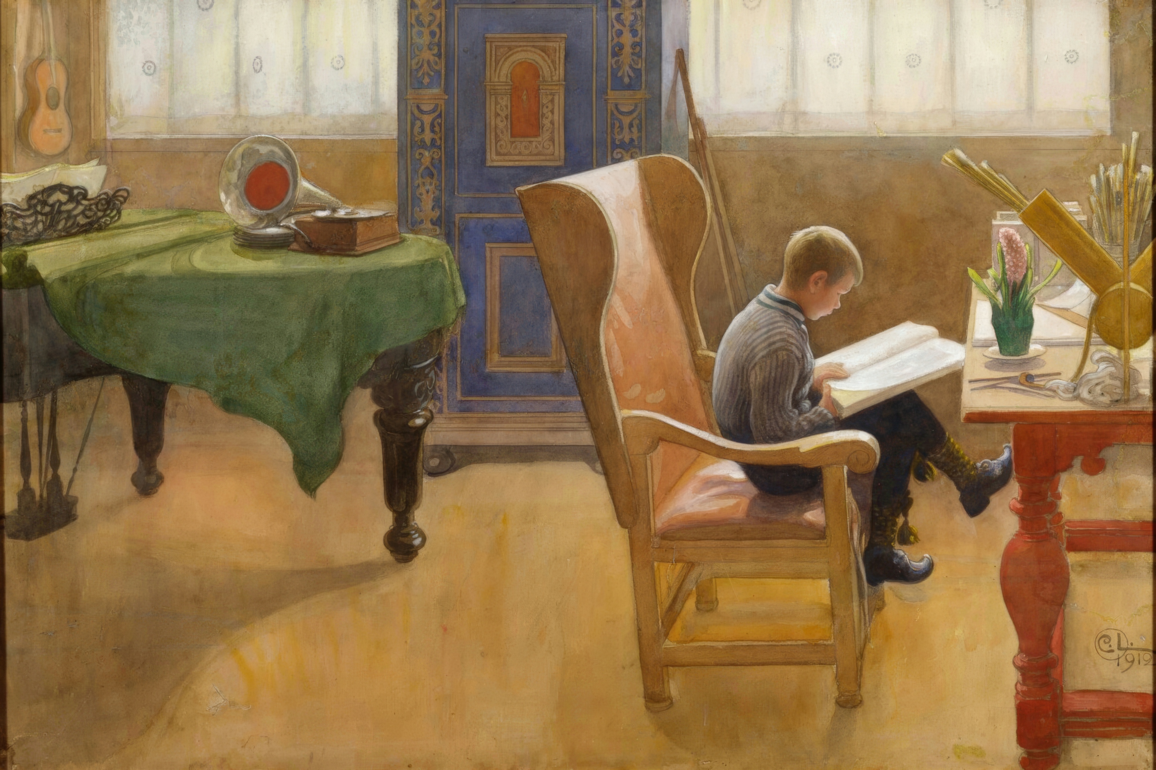 A boy reads a large book in a high backed chair at a red desk in a bright room.