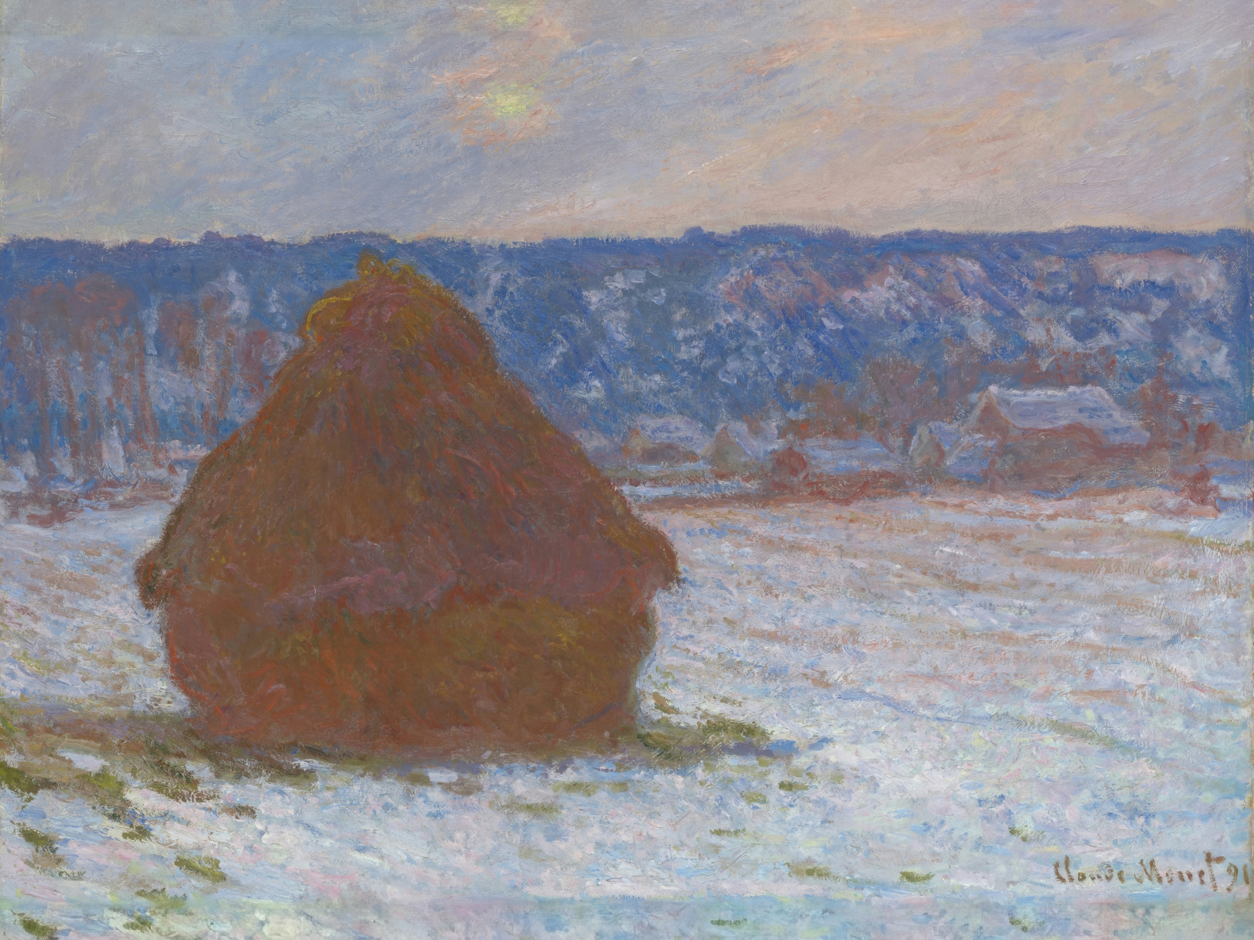 A large stack of wheat stands in a snowy field under a grey sky, with houses and trees behind.
