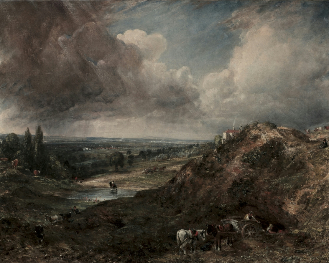 A wide view over a heath with a small pond, dark hills and a vast cloudy sky with light breaking through.