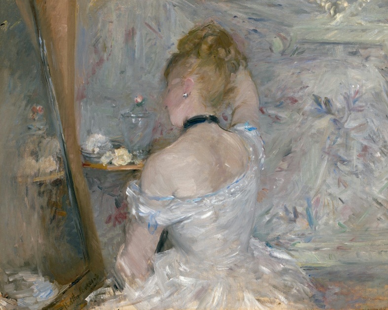 A woman in a white gown, seen from behind, sits at a dressing table before a mirror in soft light.